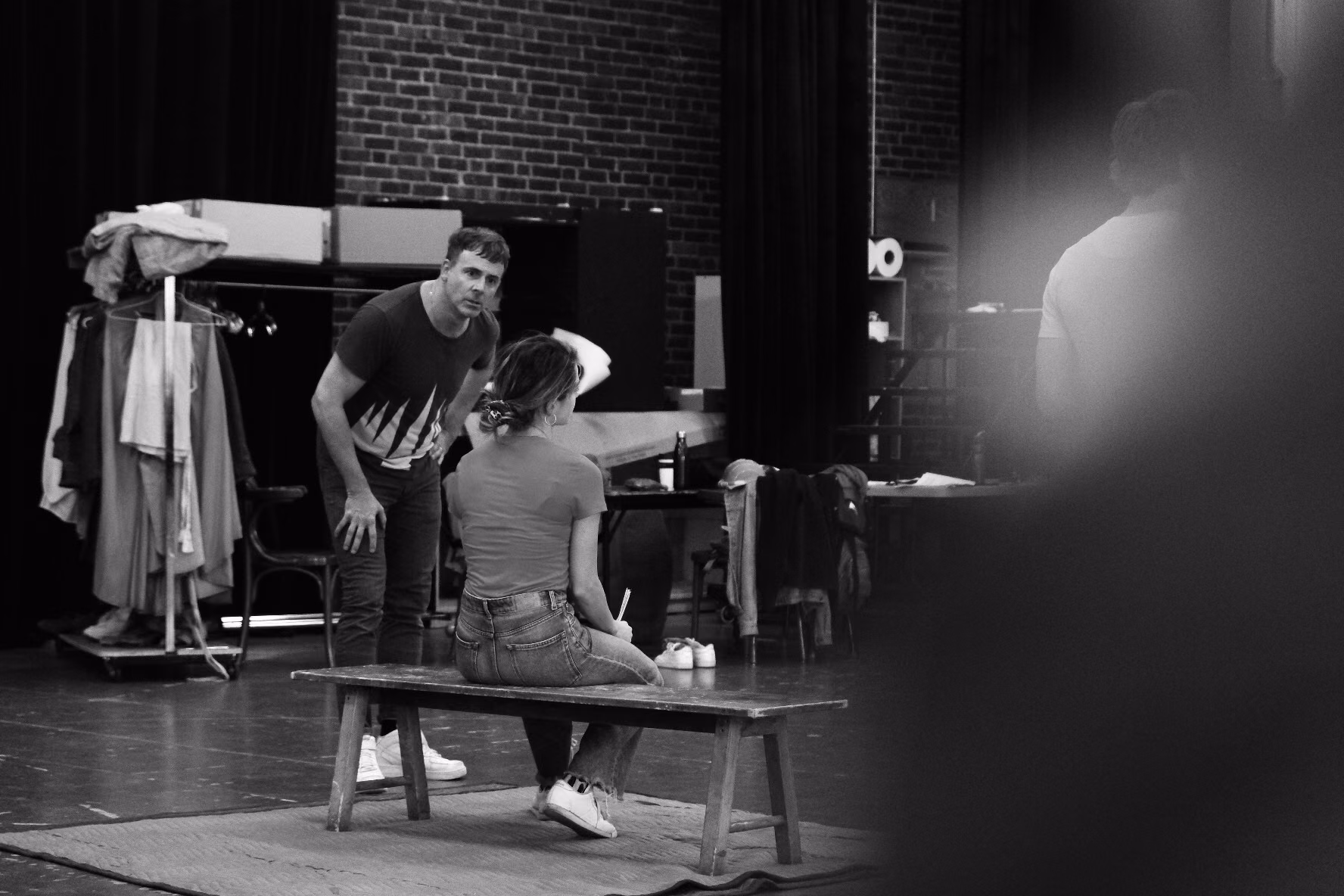 Two actors in rehearsal on a bare stage, one seated on a bench and one leaning in to speak, with a costume rack behind them.